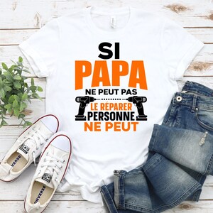 K&ouml;nnte beinhalten: Wei&szlig;es T-Shirt mit dem Text "Si Papa ne peut pas le reparer personne ne peut" in Schwarz und Orange. Der Text ist von zwei Bildern von Bohrmaschinen umgeben.