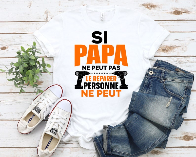K&ouml;nnte beinhalten: Wei&szlig;es T-Shirt mit dem Text "Si Papa ne peut pas le reparer personne ne peut" in Schwarz und Orange. Der Text ist von zwei Bildern von Bohrmaschinen umgeben.