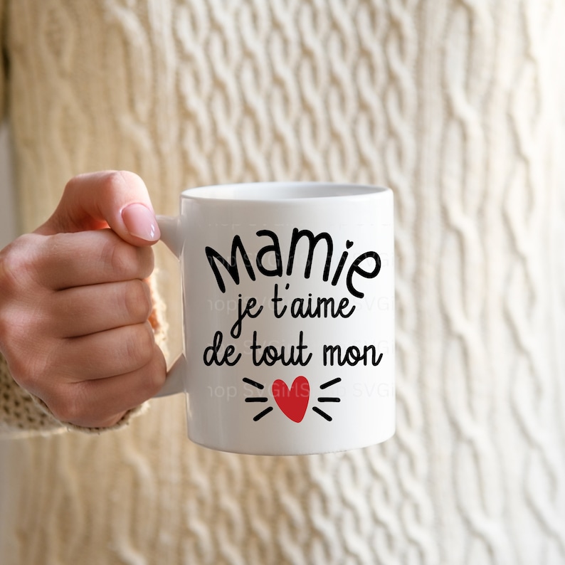 May include: White ceramic mug with a black inscription that reads "Mamie je t'aime de tout mon" with a red heart and black lines on either side of the heart.