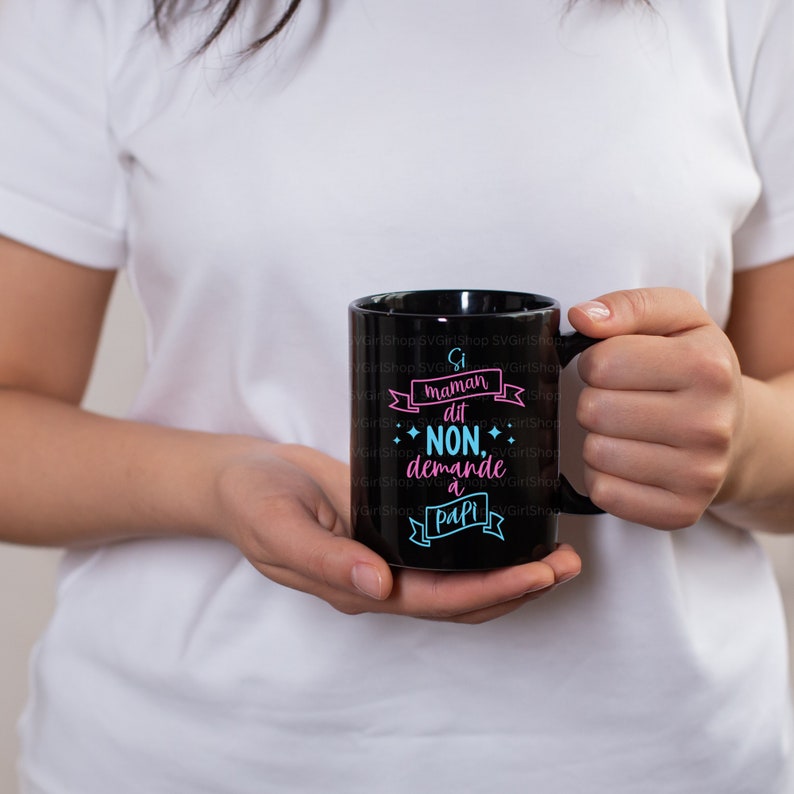 Peut inclure: Mug noir avec l'inscription "Si maman dit NON, demande &agrave; Papi" en rose et bleu. La tasse est tenue par une personne portant un t-shirt blanc. Le texte est dans un design de banni&egrave;re.