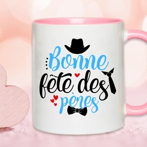 K&ouml;nnte beinhalten: Wei&szlig;e Keramiktasse mit rosafarbenem Rand und Henkel. Die Tasse hat ein blaues und schwarzes Design mit dem Text "Bonne f&ecirc;te des p&egrave;res" und einer kleinen Fliege.