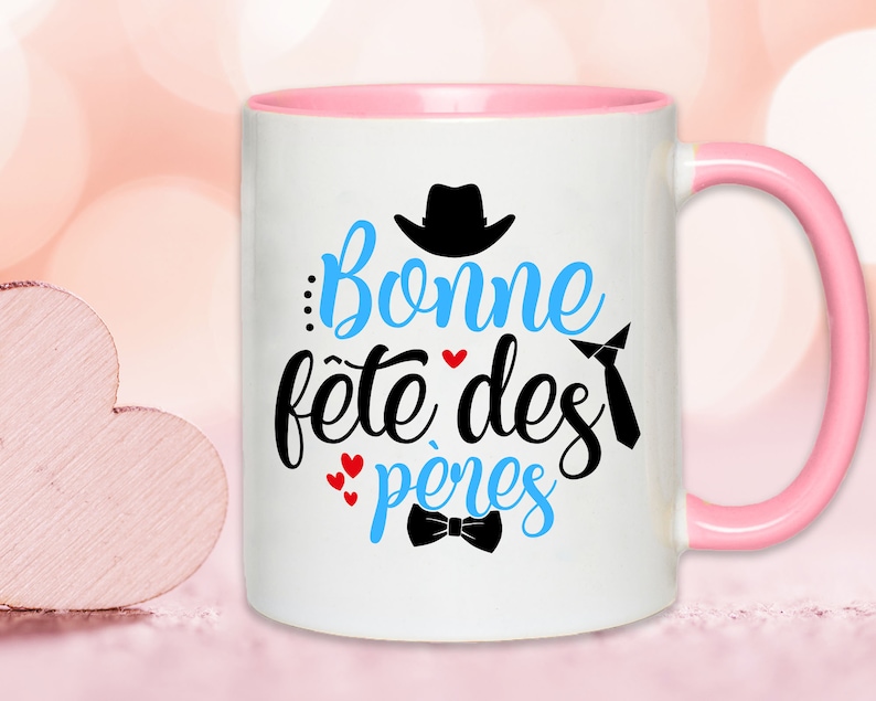 K&ouml;nnte beinhalten: Wei&szlig;e Keramiktasse mit rosafarbenem Rand und Henkel. Die Tasse hat ein blaues und schwarzes Design mit dem Text "Bonne f&ecirc;te des p&egrave;res" und einer kleinen Fliege.