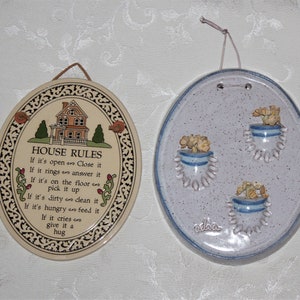May include: Two oval ceramic wall plaques. One has a house illustration and the text "HOUSE RULES". The other has three small blue planters with flowers. Both have strings for hanging.