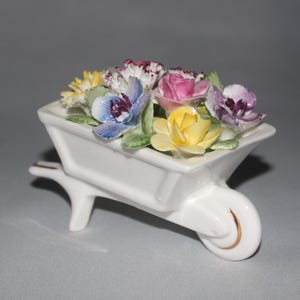 May include: A white ceramic wheelbarrow planter filled with colourful, detailed flowers. The flowers include yellow, pink, purple, and blue blooms with green leaves. The wheelbarrow has a gold accent on the wheel.