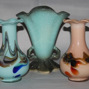 May include: Three glass vases with different designs. The vase on the left is light blue with brown and white swirls. The vase in the middle is light blue with a speckled finish and two handles. The vase on the right is light pink with brown and orange swirls.