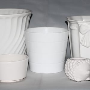 May include: Five white ceramic planters of varying sizes and shapes. The largest planter has a wavy design, while the second largest has a ribbon design. The smallest planter is shaped like a hedgehog.
