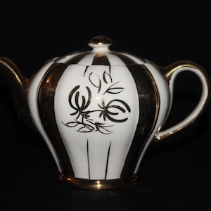 May include: A vintage teapot with a white and gold striped design. The teapot features a gold handle, spout, and base. A floral design is painted on the front. The lid is topped with a gold knob. The teapot is set against a black background.