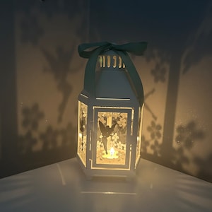 May include: White lantern with a green ribbon and a fairy silhouette design. The lantern has a candle inside and is casting a warm glow.