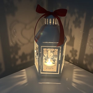 May include: White metal lantern with a red bow and a candle inside. The lantern has a floral design and casts a shadow on the white surface it is sitting on.