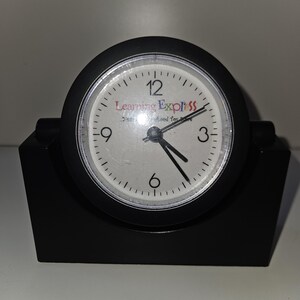 May include: A black desk clock with a white face and black hands. The clock face has black numbers and the text "Learning Express" in red, blue, and green. The clock is set on a black base.