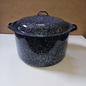 May include: A large, round, deep stock pot with a matching lid, featuring a dark navy blue enamel finish with a white speckled pattern throughout. The pot includes two side handles and a small handle on the center of the lid for easy lifting.