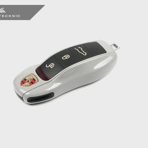 May include: A light gray car key fob with black buttons and a Porsche emblem. The fob has a sleek, modern design with keyless entry and remote start functions. The top of the fob has the text "AUTOTECKNIC".