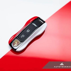 May include: A red, white, and black car key with the Porsche emblem. The key has three buttons and the text "RS". The key is on a red and white surface.