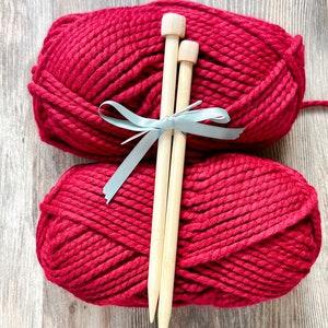 May include: Two large skeins of deep red yarn stacked vertically, secured together with a light blue ribbon. Two smooth, light-colored wooden knitting needles are placed vertically over the center of the yarn, held in place by the ribbon.