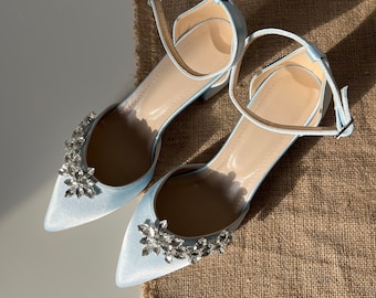 Puede incluir: Un par de bailarinas de satén azul claro con puntera y correas en el tobillo. Cada zapato está adornado con un adorno floral de cristal brillante. Los zapatos se muestran sobre una superficie texturizada de tono neutro.