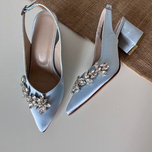 以下が含まれることがあります： 先のとがったつま先と前面にクリスタル装飾が施された、薄い青色のサテン素材のハイヒールシューズのペア。シューズには足首ストラップが付いています。