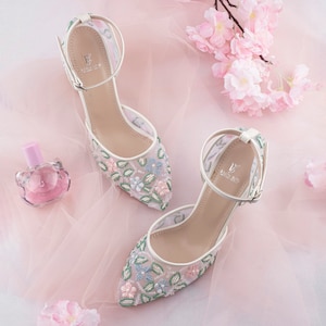 May include: A pair of white satin heels with delicate floral embroidery. The heels feature a delicate ankle strap with a small logo. The shoes are on a pink background.