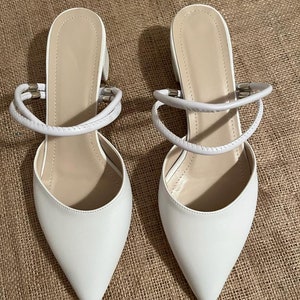 May include: A pair of white pointed-toe heeled shoes with a strap detail around the ankle.