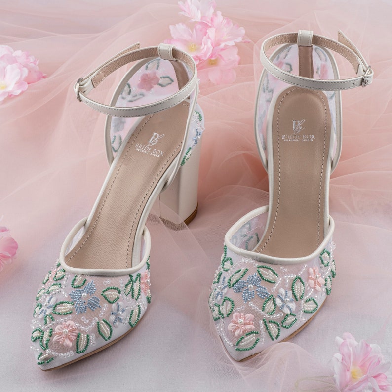 May include: White bridal heels with a delicate floral design embroidered with beads. The heels have a block heel and an ankle strap.