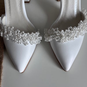 May include: A pair of white satin bridal shoes with a pointed toe and a block heel. The shoes are decorated with a pearl and crystal embellishment around the ankle.