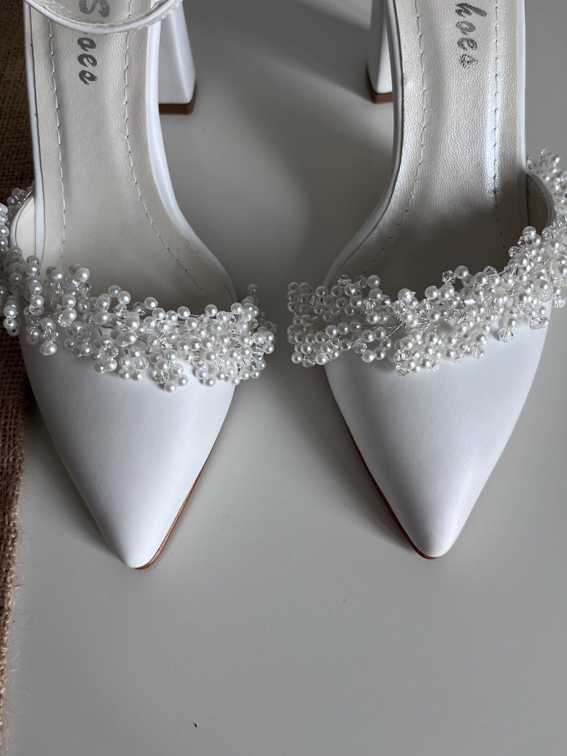 May include: A pair of white satin bridal shoes with a pointed toe and a block heel. The shoes are decorated with a pearl and crystal embellishment around the ankle.