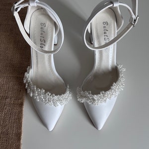 May include: White satin bridal shoes with a pointed toe and ankle strap. The shoes feature a delicate pearl embellishment around the toe.