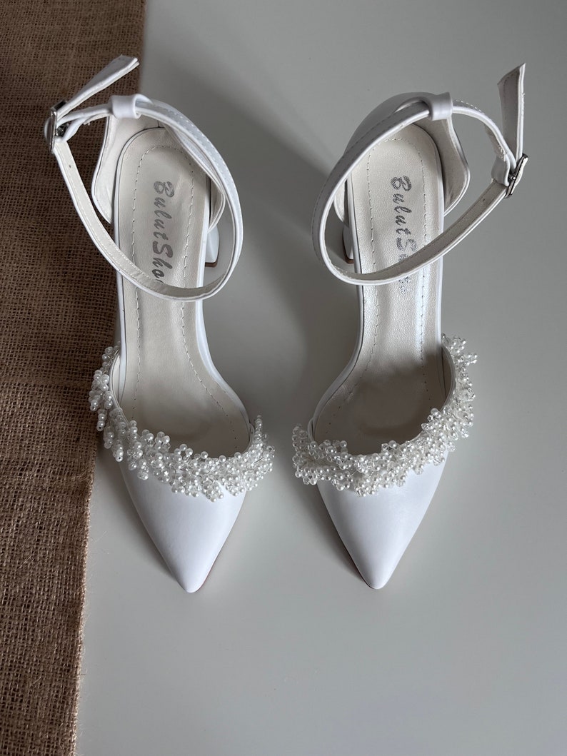 May include: White satin bridal shoes with a pointed toe and ankle strap. The shoes feature a delicate pearl embellishment around the toe.