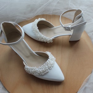 May include: A pair of white bridal shoes with pearl embellishments. The shoes have a closed toe and a high heel.