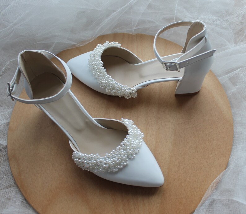 May include: A pair of white bridal shoes with pearl embellishments. The shoes have a closed toe and a high heel.