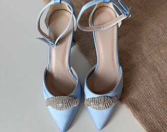 Puede incluir: Un par de zapatos de tacón de satén azul claro con correas de tobillo y un adorno de pedrería en la punta.