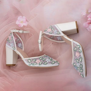 May include: White high-heeled shoes with a floral design. The shoes have a delicate mesh fabric with embroidered flowers in shades of pink, blue, and green. The heels are block heels and the shoes have ankle straps.