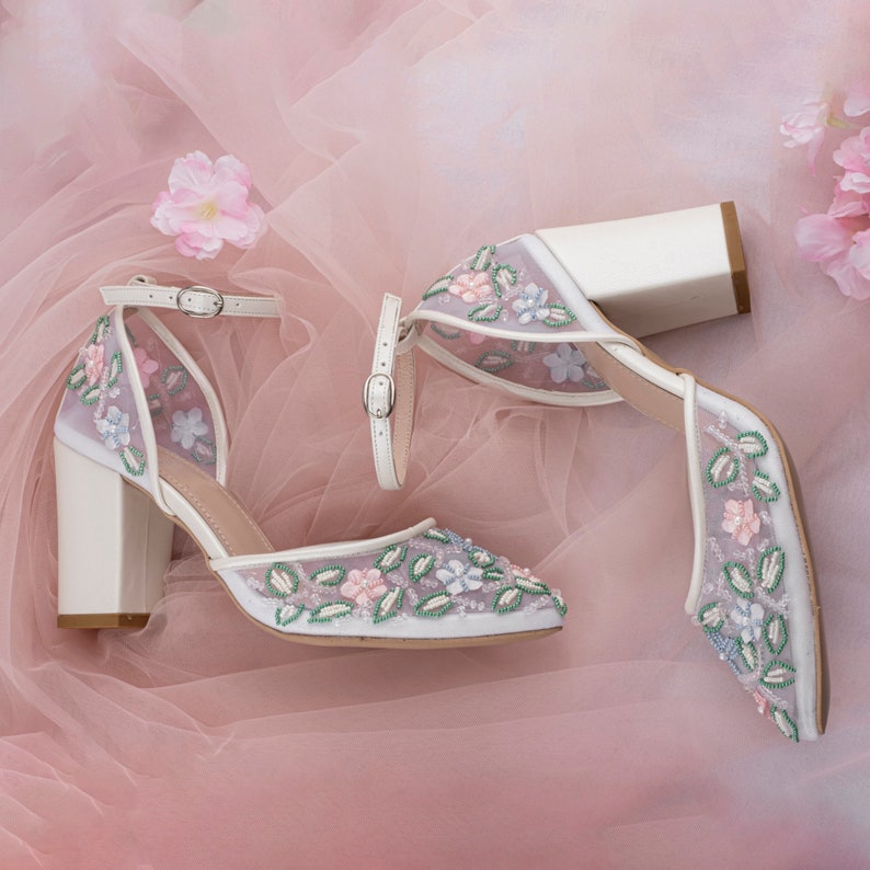 May include: White high-heeled shoes with a floral design. The shoes have a delicate mesh fabric with embroidered flowers in shades of pink, blue, and green. The heels are block heels and the shoes have ankle straps.