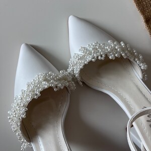 May include: A pair of white satin bridal shoes with pearl embellishments. The shoes have a pointed toe and an ankle strap.