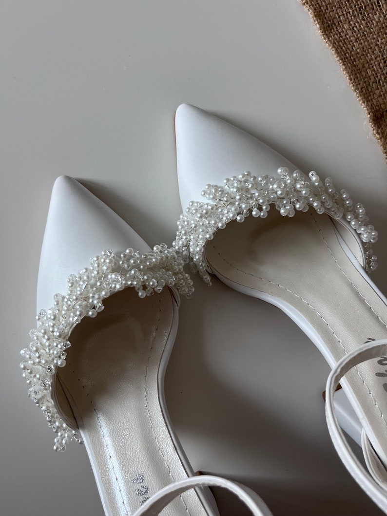 May include: A pair of white satin bridal shoes with pearl embellishments. The shoes have a pointed toe and an ankle strap.
