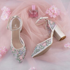 May include: A pair of white high-heeled shoes with floral embroidery. The shoes have a delicate ankle strap and a block heel. The shoes are on a pink tulle background with pink flowers.