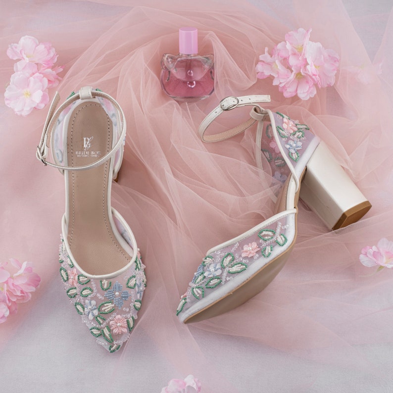 May include: A pair of white high-heeled shoes with floral embroidery. The shoes have a delicate ankle strap and a block heel. The shoes are on a pink tulle background with pink flowers.