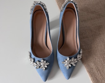 Puede incluir: Un par de zapatos de tacón de aguja de ante azul claro con punta puntiaguda y un adorno de pedrería en la parte delantera de cada zapato.