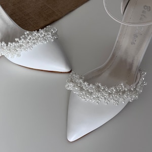 May include: A pair of white bridal shoes with a pointed toe and a delicate pearl embellishment around the ankle strap.