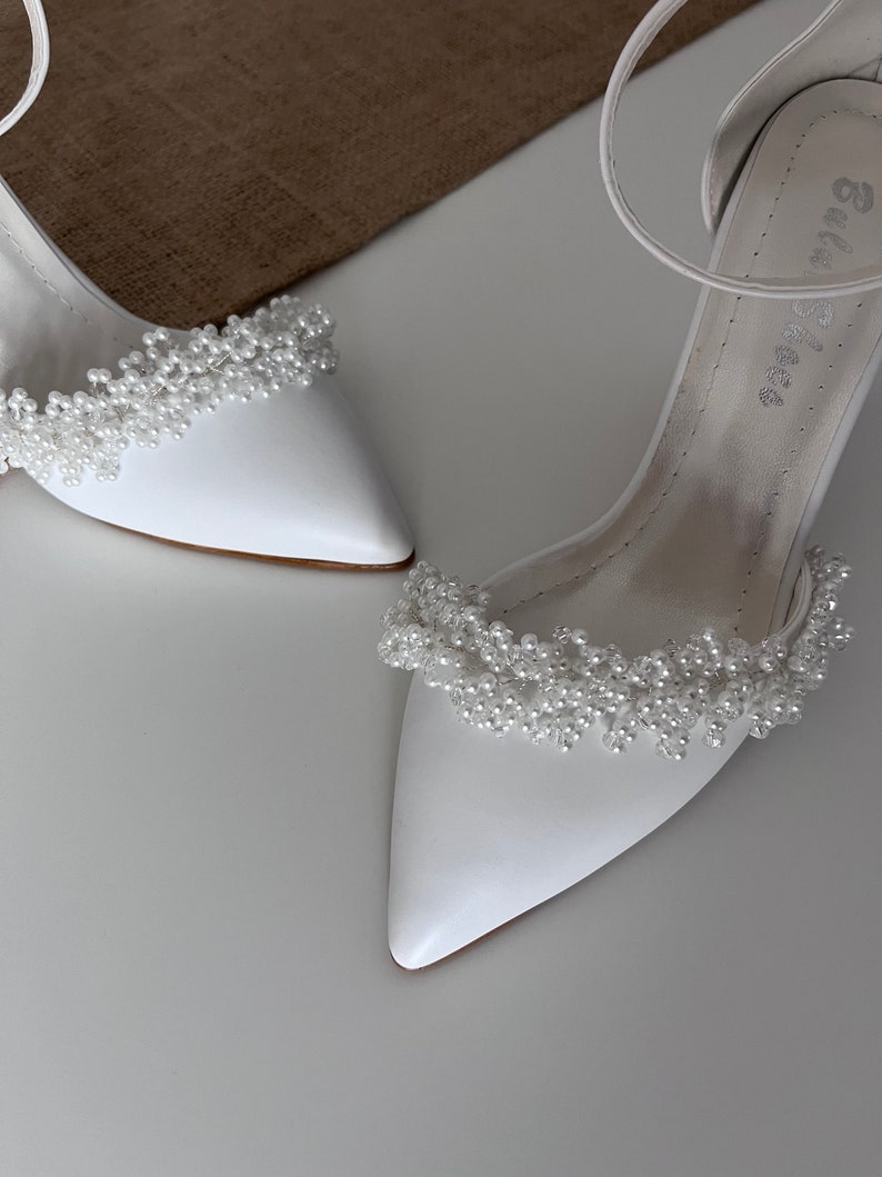 May include: A pair of white bridal shoes with a pointed toe and a delicate pearl embellishment around the ankle strap.