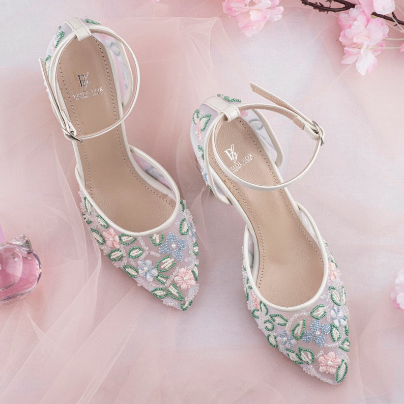 May include: A pair of white bridal shoes with delicate floral embroidery in shades of pink, blue, and green. The shoes have a delicate ankle strap with a buckle closure. The brand name "BRIDAL BOX" is visible on the insole.