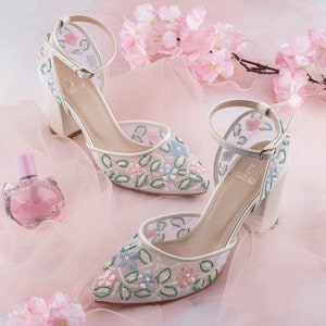 May include: A pair of white high-heeled shoes with delicate floral embroidery. The shoes have a mesh upper and a pointed toe. The embroidery features pink, blue, and green flowers.