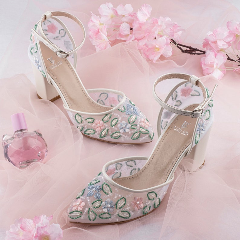 May include: A pair of white high-heeled shoes with delicate floral embroidery. The shoes have a mesh upper and a pointed toe. The embroidery features pink, blue, and green flowers.