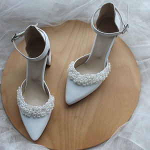May include: A pair of white satin bridal shoes with pearl embellishments on the toe. The shoes are on a wooden surface.