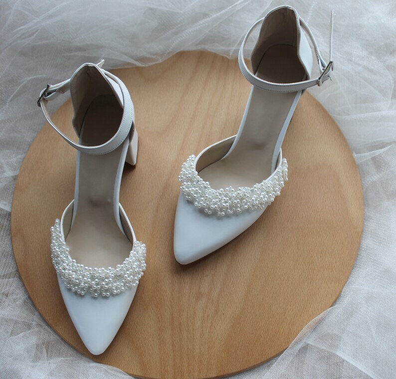 May include: A pair of white satin bridal shoes with pearl embellishments on the toe. The shoes are on a wooden surface.