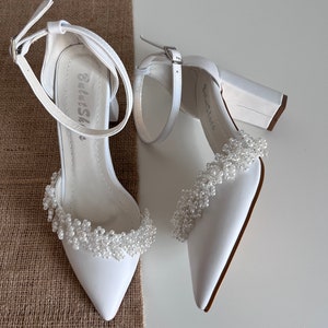 May include: A pair of white satin bridal heels with a block heel and a beaded embellishment around the ankle strap. The heels have a pointed toe and a buckle closure.