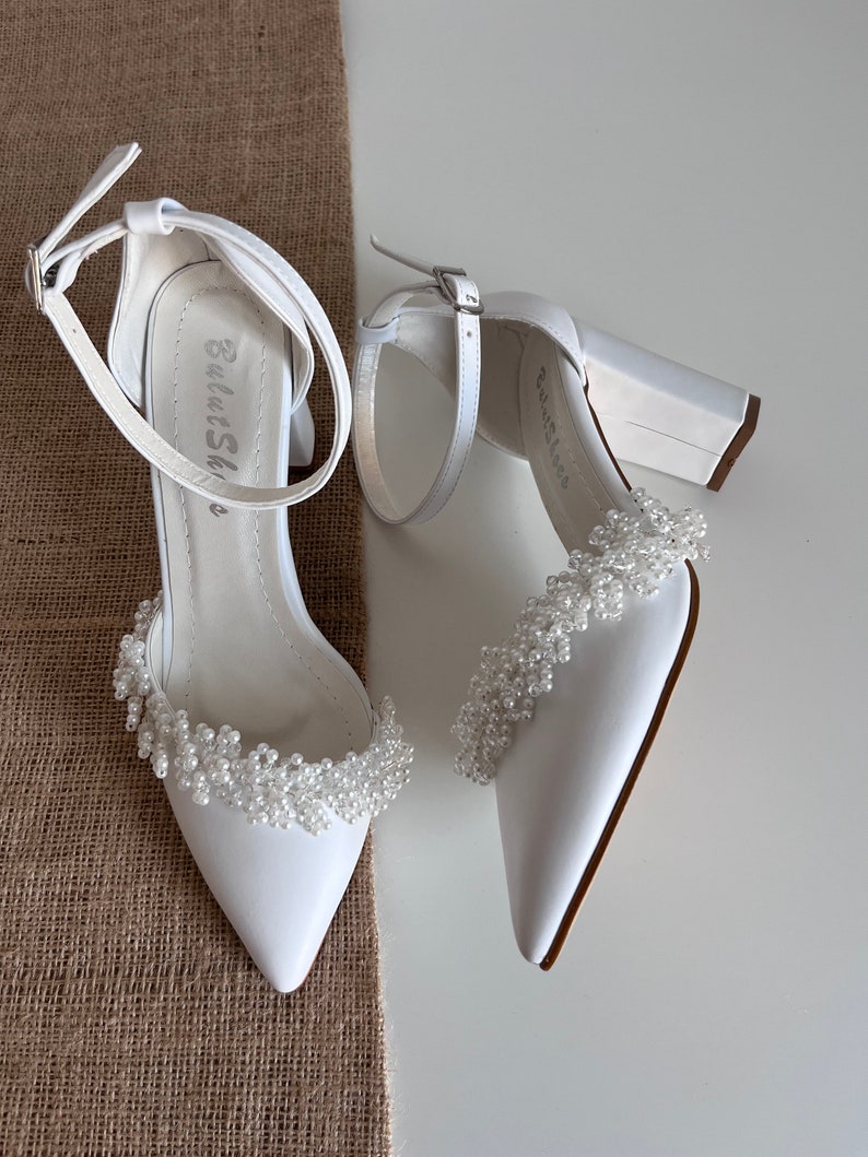 May include: A pair of white satin bridal heels with a block heel and a beaded embellishment around the ankle strap. The heels have a pointed toe and a buckle closure.