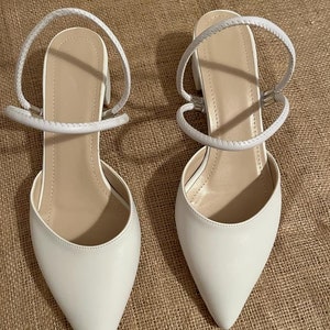 May include: A pair of white pointed-toe heeled sandals with ankle straps. The straps are made of a braided material.