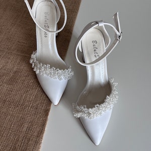 May include: A pair of white satin bridal shoes with a pointed toe and ankle strap. The shoes are decorated with a row of white pearl beads around the ankle strap.