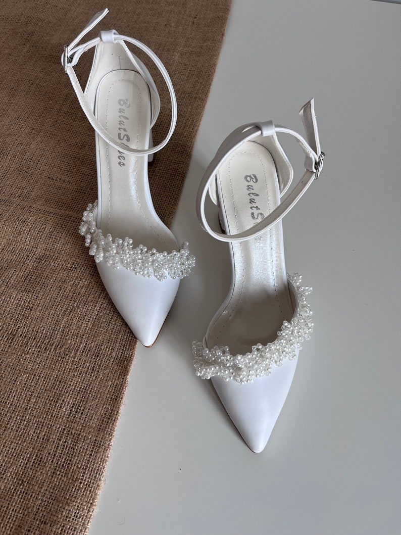 May include: A pair of white satin bridal shoes with a pointed toe and ankle strap. The shoes are decorated with a row of white pearl beads around the ankle strap.