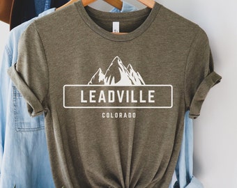 Op de afbeelding: Een groen T-shirt met een witte afbeelding van bergen en de tekst "Leadville, Colorado".